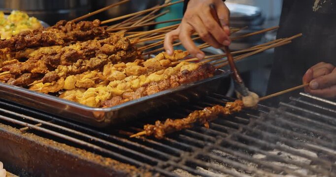 Chef brushing oil on meat skewers over charcoal grill, traditional Asian street food market, outdoor cooking process, culinary documentary.