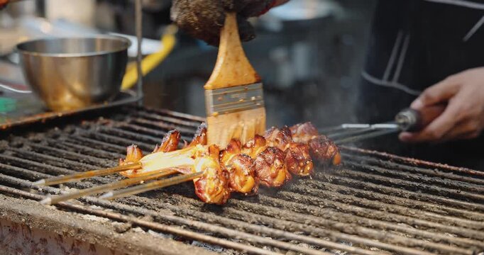 Chef brushing sauce on meat skewers, grilling on outdoor street food stall, Asian night market culinary experience, professional cooking process.