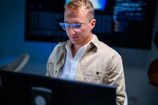 A light-haired cyber defence engineer watches live network activity on his monitor inside a modern security operations centre after hours, protecting critical data.