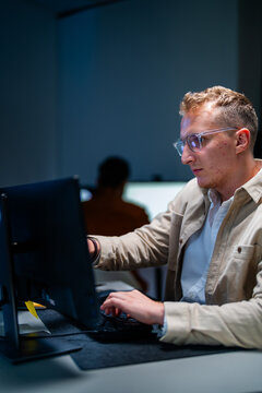 A blond male network security analyst enters commands at his workstation in a shadowy data monitoring room lit by screen glow, during an overnight monitoring shift.