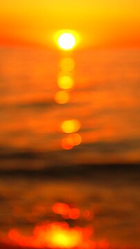 Vibrant ocean sunset bokeh illuminates rolling sea waves. Vertical orientation captures shifting golden light reflections on the water surface in a tranquil scene.