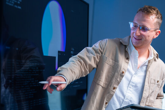 A light-haired cyber defence engineer stands holding a tablet while briefing a colleague inside a dark security operations centre at night, under the blue glow of security dashboards.