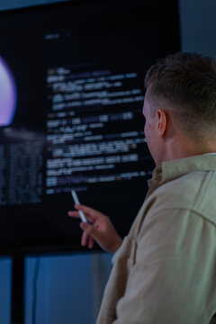 A young bearded security engineer reaches toward the screen to highlight data in a dimly lit network monitoring room, reviewing code and system logs for signs of a cyber attack.