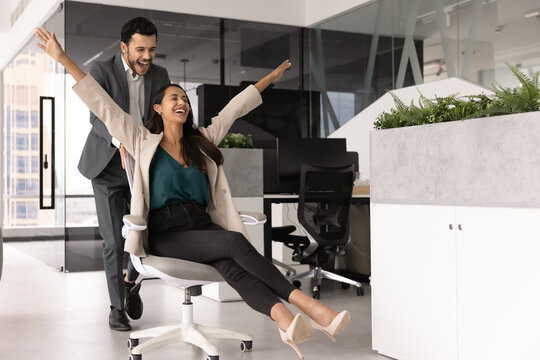 Two laughing multiethnic coworkers celebrating achievements, project or workday completion, riding playfully on office chairs at workplace. Amusement, fun during break, victory, corporate success