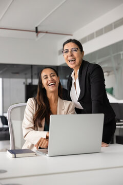 Diverse businesswomen, team members laughing, look at camera, working on project or digital tasks in corporate office, represents successful collaborative teamwork, business achievements at workplace