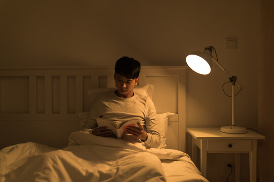 Young man reading a book in bed at night under bedside lamp