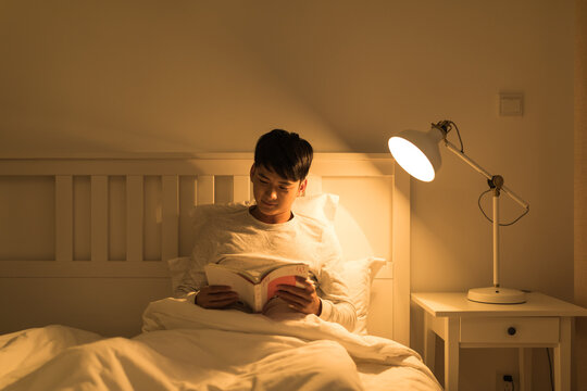 Young man reading a book in bed at night in a cozy bedroom