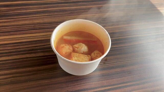Hot spicy tom yum soup served in a white paper bowl with fish cakes, crab stick, and meatballs on a wooden table.