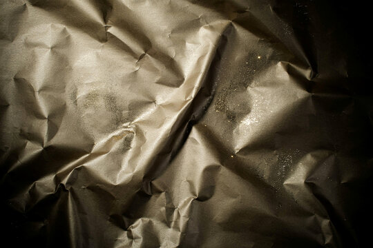 Abstract close-up of a heavily crumpled and creased metallic golden brown paper surface covered in scattered sparkling glitter particles with strong contrasting shadows and highlights