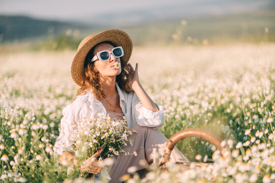 Relaxed woman holding Matricaria chamomilla bouquet while sitting in a sunny spring meadow with a picnic basket