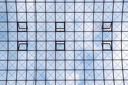 Blick von unten auf eine moderne Glasdachkonstruktion mit quadratischen Fenstern und geometrischem Stahlgitter. Der helle Himmel schimmert durch die gro&szlig;en Scheiben.