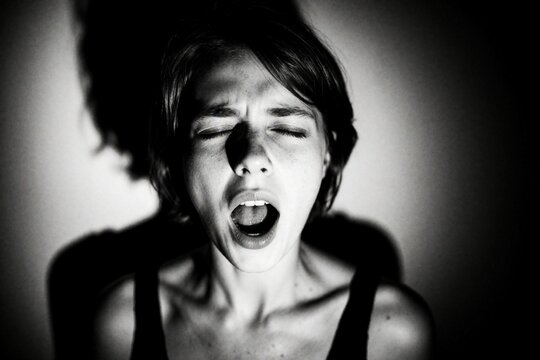 Woman screams with eyes closed in darkness. Intense facial expression conveys raw emotion, pain, or catharsis. Dramatic BW portrait captures inner struggle and vulnerability.