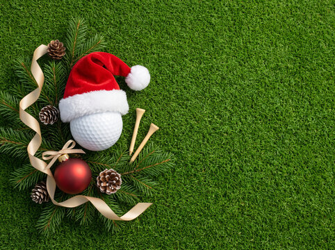 Christmas golf flat lay photographed directly overhead on lush, freshly manicured green golf grass. On the left side, one large plain white golf ball wearing a luxurious red Santa hat with soft fluffy