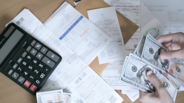 Top view of hands counting US dollar banknotes with calculator, bills and financial documents on desk, concept of household expenses, budgeting, debt, payment and money management.