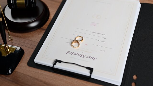 Official marriage certificate lies over wooden desk, facilitating marriage registration paperwork inside court setting, marriage license approval requiring valid certificate status beside judge gavel.