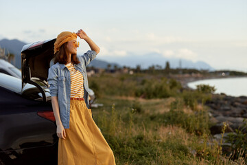 Naklejka premium Woman outdoors by a car on a coastal landscape, wearing a denim jacket and mustard dress, sunlit moment, joyful pose, gentle breeze, vibrant colors, relaxed lifestyle