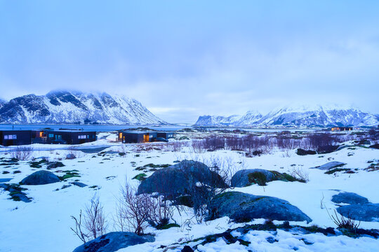 Serene Winter Twilight in Lofoten Norway: Modern Dark Cabins with Warm Glowing Lights Amidst Snow-Covered Landscape and Majestic Arctic Mountains, Moody Blue Hour Nordic Travel Vista.