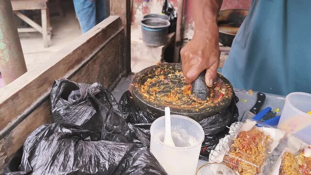 BANDUNG, INDONESIA - AUGUST 19, 2026: Indonesian street food vendor grinding spicy chili sauce with a traditional stone mortar and pestle at a local food stall