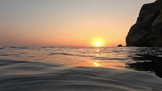 Golden sunset glowing over tranquil sea waves beside a rocky cliff. Low angle tracking shot.