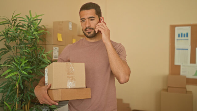 Man carrying stacked cardboard boxes and talking on phone while smiling inside a building; small business optimism.