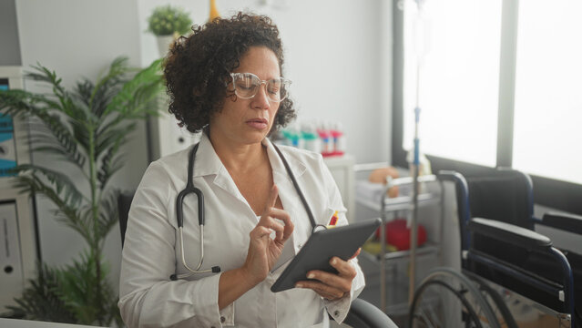 Woman doctor in white coat and stethoscope taps tablet with fingers, wearing glasses beside wheelchair and medical cart in building; care and focus.