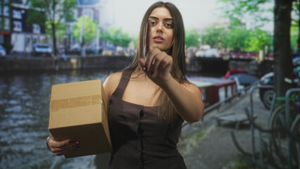 Fototapeta premium Woman holding cardboard box points index finger on street by a canal and bicycles; determination delivery.