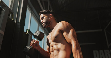 Fototapeta premium Dumbbells, exercise and bodybuilder man with music and serious for training, muscle growth and self care. Earphones, workout playlist and person or low angle in gym for fitness, strong and power