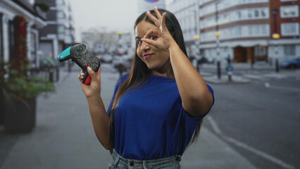 Young woman holding a gamepad while smiling and making an ok sign over her eye on a street with blurred buildings and parked cars  playful fun. © Krakenimages.com