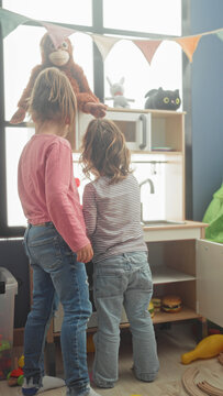 Girl and younger child play at a wooden toy kitchen, holding plates and a stuffed monkey beside a window with bunting in a building; joy.