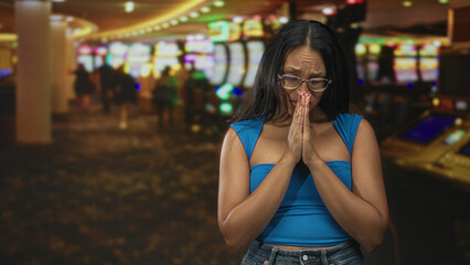 Fototapeta premium Woman with hands pressed together in prayer in a casino with slot machines and blurred patrons; desperation hope loss.