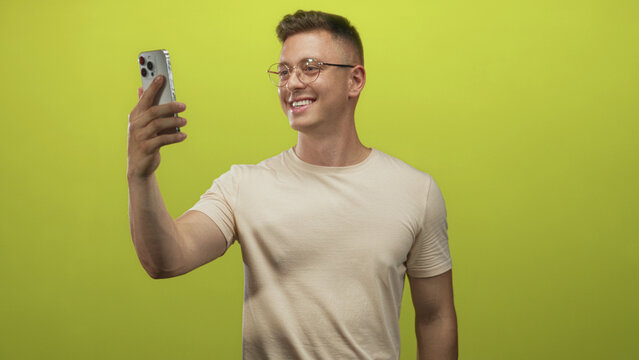 Man taking selfie with smartphone, arm extended showing hand and forearm, wearing glasses in studio setting with lime wall; casual confidence.