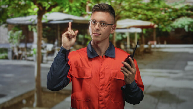 Young blond man in orange coverall holding a walkietalkie and points finger up on a street cafe terrace while wearing glasses and looking focused; alert readiness.