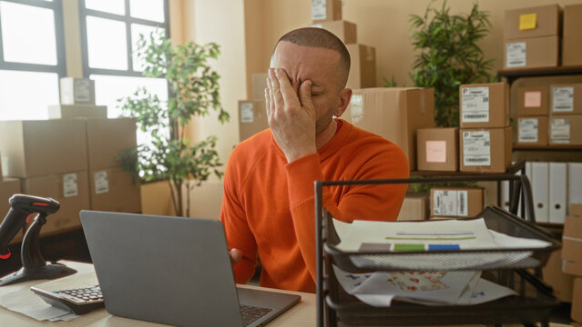 Man writing with pen at laptop and cardboard boxes, hand to forehead from fatigue in building; small business stress.
