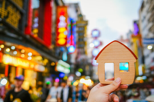 Real estate and urban property concept with hand holding wooden house model against blurred night market street with glowing neon lights for city apartment, commercial shop and downtown home.