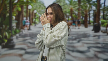Fototapeta premium Woman with hands covering face on a palm lined street, wearing striped shirt and jeans, side profile visible; anxiety vulnerability.