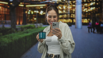 Fototapeta premium Woman holding smartphone and teal mug, smiling while reading a message on her phone outside illuminated building entrance; joy connection.