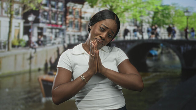 Woman wearing white polo shirt with hands clasped in prayer and pleading facial expression on a street beside a canal bridge; pleading vulnerability.