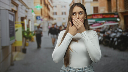 Fototapeta premium Woman covering mouth with hands and long hair on a crowded street with scooters and shops; surprise reaction.