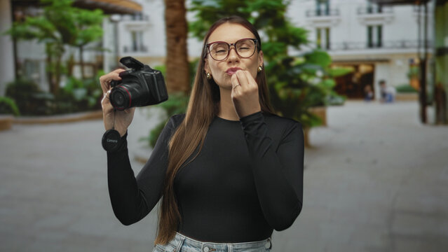 Woman holding dslr camera pinches fingers to lips while touching nose and gesturing, posing in a street courtyard with buildings and palm trees; playful tutorial.