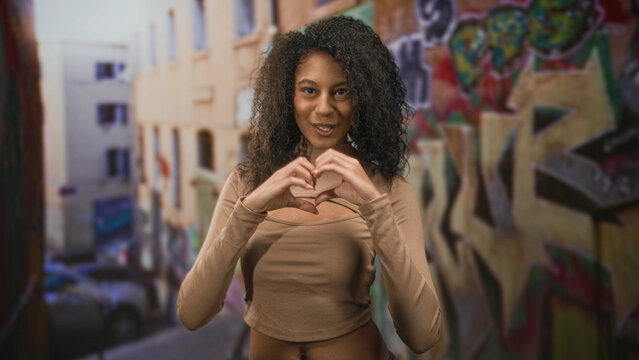 Teenage woman makes a heart with her hands and smiles, exposed midriff visible, standing by graffiti covered wall on a street; joyful self love.