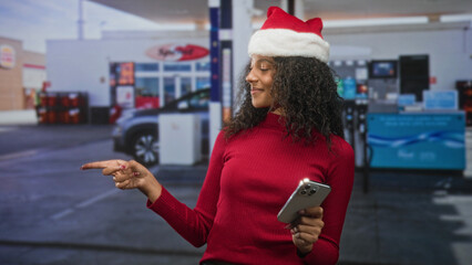 Woman wearing santa hat holds smartphone and points finger while smiling in building entrance  holiday cheerful. © Krakenimages.com