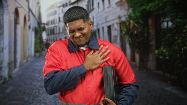 Man in uniform clutching chest holding toolbox on cobbled street for heartattack; medical emergency distress.