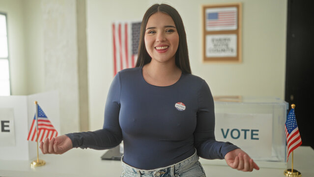Woman wearing a voted sticker gestures with open hands at a polling booth with us flags inside a voting building; civic pride.