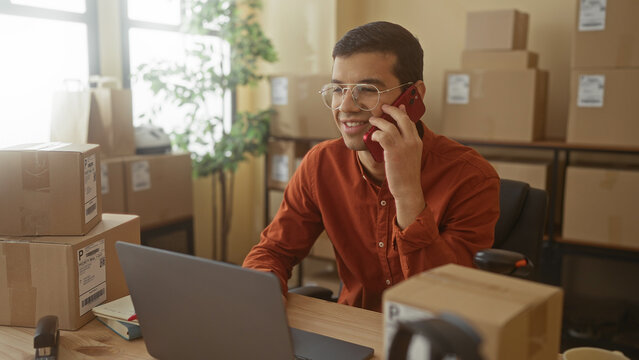 Man holding smartphone to ear and typing on laptop with visible hand among shipping boxes in building; entrepreneurship focused.