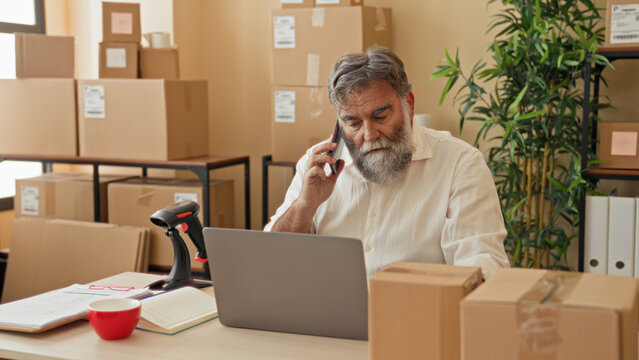 Man holding phone to ear and typing on laptop amid cardboard boxes and barcode scanner in building; small business focus.