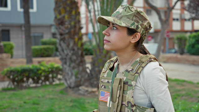 Woman soldier in army uniform and camouflage cap with us flag patch looks up in profile, face visible on street near palm trees and building; pride duty.