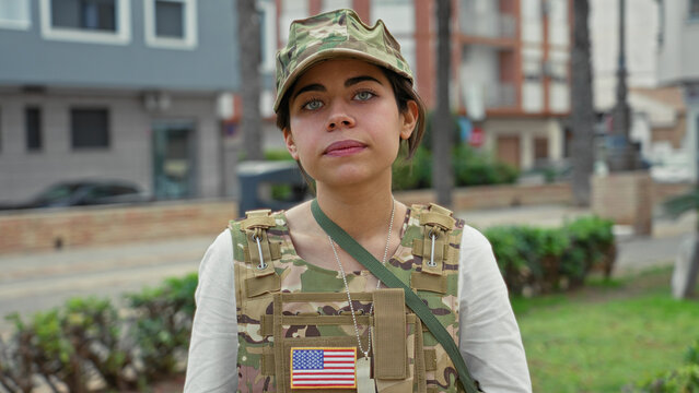 Woman soldier wearing camouflage cap and tactical vest with us flag patch and dog tags looks at camera on street near low wall and palms; pride duty.