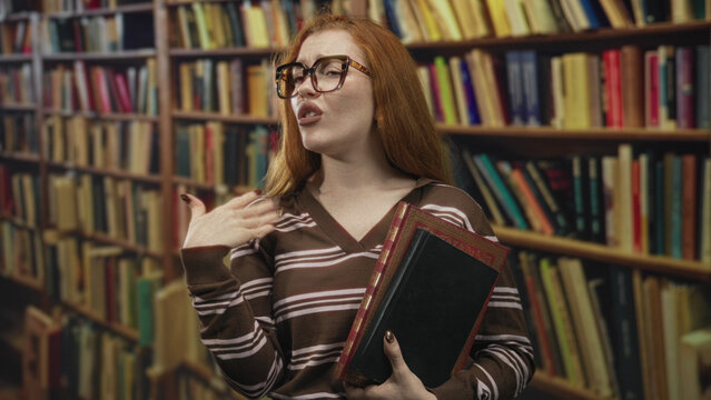 Redhead woman holding a hardcover book with hand lifted in a dismiss gesture in library; smug confidence.