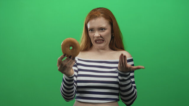 Woman recoils and pushes a glazed donut away with her hand in a green studio, making a disgusted face; disgust.