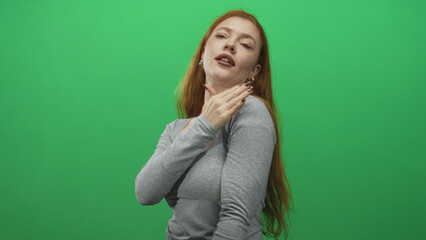 Fototapeta premium Redhead woman flipping hair and touching neck in a green screen studio, wearing gray top and hoop earrings; confidence.